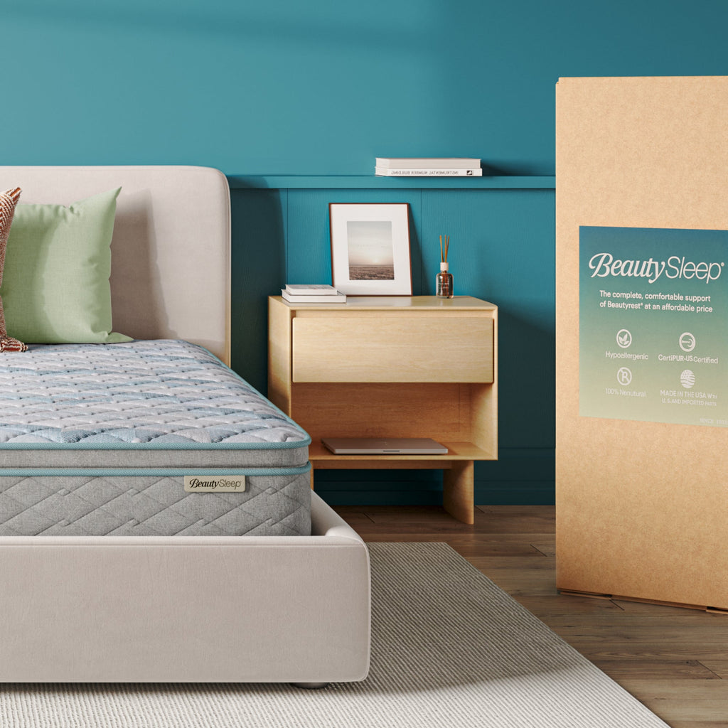 Beautysleep Mattress in a grey bedframe with 3 pillows on top of it within a bedroom setting with BeautySleep packaging to the right of it.